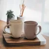 Two ceramic mugs with a textured diamond pattern are placed on a wooden cutting board, with a small potted plant and a diffuser in the background. Produktbild-Vorschau