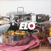 A collection of ELO brand cookware, including pots and a frying pan with food, arranged on a wooden surface with a red and white checkered cloth. Náhled obrázku produktu