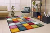 A colorful, modern rug with a checkered design is laid out on a wooden floor in a living room. The rug is placed in front of a sofa and a chair, with a bookshelf in the background. Produktbild-Vorschau