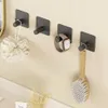 Four black adhesive hooks are mounted on a light beige wall. A loofah is hanging from the first hook, an Apple Watch from the second, and a wooden-bristled brush from the fourth. The third hook is empty. A white countertop is visible in the foreground. Anteprima dell’immagine prodotto