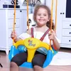 A young girl is sitting on a colorful swing indoors, with furniture in the background. Produktbild-Vorschau