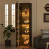 A tall, narrow display cabinet with glass doors and shelves illuminated by warm LED lights, showcasing several decorative busts and figurines. The cabinet is positioned in a room next to a doorway and a large potted plant, with a sofa visible in the backgr Produktbild-Vorschau