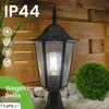 A black outdoor lantern with a light bulb inside, set against a wooden fence and white stones. Rain is depicted falling. Produktbild-Vorschau