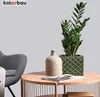 A green ceramic planter with a geometric pattern sits on a wooden coffee table next to a beige vase. A gray armchair is visible in the background. Produktbild-Vorschau