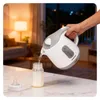A hand is pouring hot water from a white and gray electric kettle into a baby bottle on a marble countertop. The background shows a softly lit living room with a couch and lamp. Produktbild-Vorschau