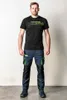 A man stands facing the camera wearing a black t-shirt and blue work pants with green accents. Produktbild-Vorschau