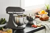A KitchenAid stand mixer is shown on a kitchen counter with various baking ingredients and a loaf of bread. Produktbild-Vorschau
