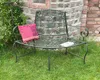 A green metal garden bench with a decorative floral backrest sits on a grassy lawn in front of a stone wall. A pink and red patterned cushion is on the left side of the bench, and a small book is on the right. Anteprima dell’immagine prodotto