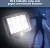 A solar-powered spotlight with a motion sensor is illuminated, casting a bright light on a shadowy figure in the background, suggesting its use for security. Produktbild-Vorschau