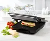 A panini press with a sandwich inside, along with some fresh ingredients like tomatoes and greens, on a kitchen counter. Podgląd zdjęcia produktu