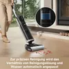 A person is using a wet and dry vacuum cleaner on a wooden floor, cleaning up a red spill. A close-up inset shows the water tank of the vacuum. Produktbild-Vorschau