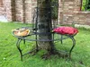A round metal bench is placed around a tree in a garden setting. A straw hat and a red patterned pillow are on the bench. Náhled obrázku produktu