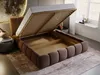 A bed with its storage compartment open, showing the slatted base and the interior storage space. The bed is upholstered in brown fabric and is placed on a rug in a room with a window and some decorative items. Produktbild-Vorschau