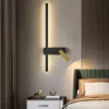 A modern wall lamp with a black vertical bar and a gold adjustable spotlight is mounted on a wall above a nightstand next to a bed. Anteprima dell’immagine prodotto
