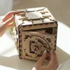 A wooden puzzle safe with its door open, showing intricate gears and a dial. Hands are interacting with the safe, suggesting assembly or operation. Produktbild-Vorschau