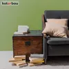 A vintage wooden chest with books on top and beside it, next to a gray armchair with beige pillows, against a light green wall. Náhled obrázku produktu