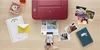 A red Canon PIXMA TS3352 printer is shown on a wooden desk, printing a document. Several printed photos, a notebook, a letter, and an envelope are scattered around the printer. A smartphone is being held by a person, displaying an image that appears to be  Anteprima dell’immagine prodotto