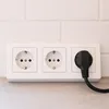 A white triple electrical outlet with a black plug inserted into the rightmost socket, set against a white tiled wall and a light gray countertop. Produktbild-Vorschau