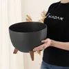 A person is holding a black planter with wooden legs. Náhled obrázku produktu