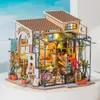 A miniature dollhouse flower shop with a greenhouse attached, illuminated by small lights. Náhled obrázku produktu