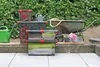 Outdoor storage boxes and gardening tools arranged on a patio with a hedge in the background. Produktbild-Vorschau