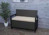 A grey rattan storage bench with a beige cushion sits on a wooden floor in front of a wood-paneled wall. A potted plant is to the left of the bench. Voir les images du produit