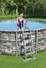 A woman is standing in an above-ground pool, holding onto a ladder. The pool has a stone-like exterior. Produktbild-Vorschau