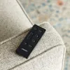 A black Bose remote control rests on the armrest of a textured, light-colored armchair. A blurred rug with a pattern of blue and orange is visible in the background. Anteprima dell’immagine prodotto