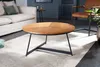 A round coffee table with a wooden top and black metal legs is placed on a rug in a living room setting. A sofa and a chair are visible in the background. Produktbild-Vorschau