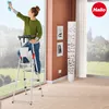 A woman is cleaning a window while standing on a stepladder indoors. Produktbild-Vorschau