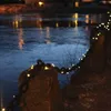 A chain decorated with fairy lights is draped over rocks next to a body of water at night. Produktbild-Vorschau