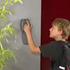 A young boy with a backpack is using a key safe mounted on a wall next to a red door. Produktbild-Vorschau