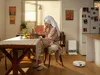 A woman in a robe and towel is eating at a table while a robot vacuum cleaner is in the background. Produktbild-Vorschau
