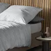 A close-up view of a bed with gray bedding and a wooden headboard. Produktbild-Vorschau