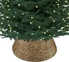 A close-up view of the bottom portion of a decorated Christmas tree, featuring green branches, warm white lights, and a woven tree skirt. Produktbild-Vorschau