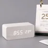 A digital alarm clock with a white wood finish is displayed on a gray surface next to a desk calendar. Produktbild-Vorschau