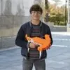A young person is holding an orange toy crossbow outdoors. Produktbild-Vorschau