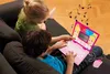 Two children are sitting on a couch, looking at a pink toy laptop with Disney Princess characters on the screen. Musical notes are depicted floating above the laptop. Produktbild-Vorschau