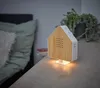 A house-shaped sound machine sits on a wooden nightstand next to a bed. Produktbild-Vorschau