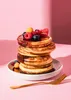 A stack of pancakes topped with berries and syrup on a white plate, with a gold fork in front of it, against a pink background with shadows. Produktbild-Vorschau
