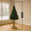 A tall artificial Christmas tree with a real wood trunk and a jute sack base, standing in a room with a large window showing a snowy outdoor scene. Produktbild-Vorschau