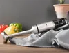 A silver and black immersion blender rests on a gray waffle-weave towel, with red, yellow, and green bell peppers in the background. A terracotta pot with a plant is visible in the upper right corner. Produktbild-Vorschau