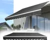 A dark gray awning is shown in two different views: one extended over a modern patio with large windows and another folded flat below. The background includes architectural elements and a hint of a residential roofline. Náhľad obrázku produktu