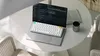 A laptop with a mechanical keyboard on top, placed on a round white table next to a newspaper and a cup. Produktbild-Vorschau