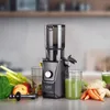 A slow juicer is shown on a countertop with fresh fruits and vegetables, and glasses of juice. Náhled obrázku produktu