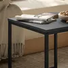 A close-up of a small black side table with a folded newspaper and a wallet on top, with a blanket and a bed in the background. Podgląd zdjęcia produktu