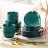 A person is holding a plate from a teal colored dinnerware set, with stacks of plates and bowls in the background. Produktbild-Vorschau