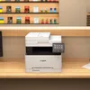 A white Canon printer is placed on a wooden desk, with shelves of products in the background. Produktbild-Vorschau