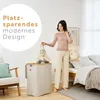 A woman is putting laundry into a tall, narrow laundry basket in a bedroom setting. Produktbild-Vorschau