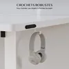 A close-up view of the underside of a white desk, showing a black hook and a pair of gray headphones hanging from it. Text overlays suggest the hook is robust and keeps items within reach. Voir les images du produit